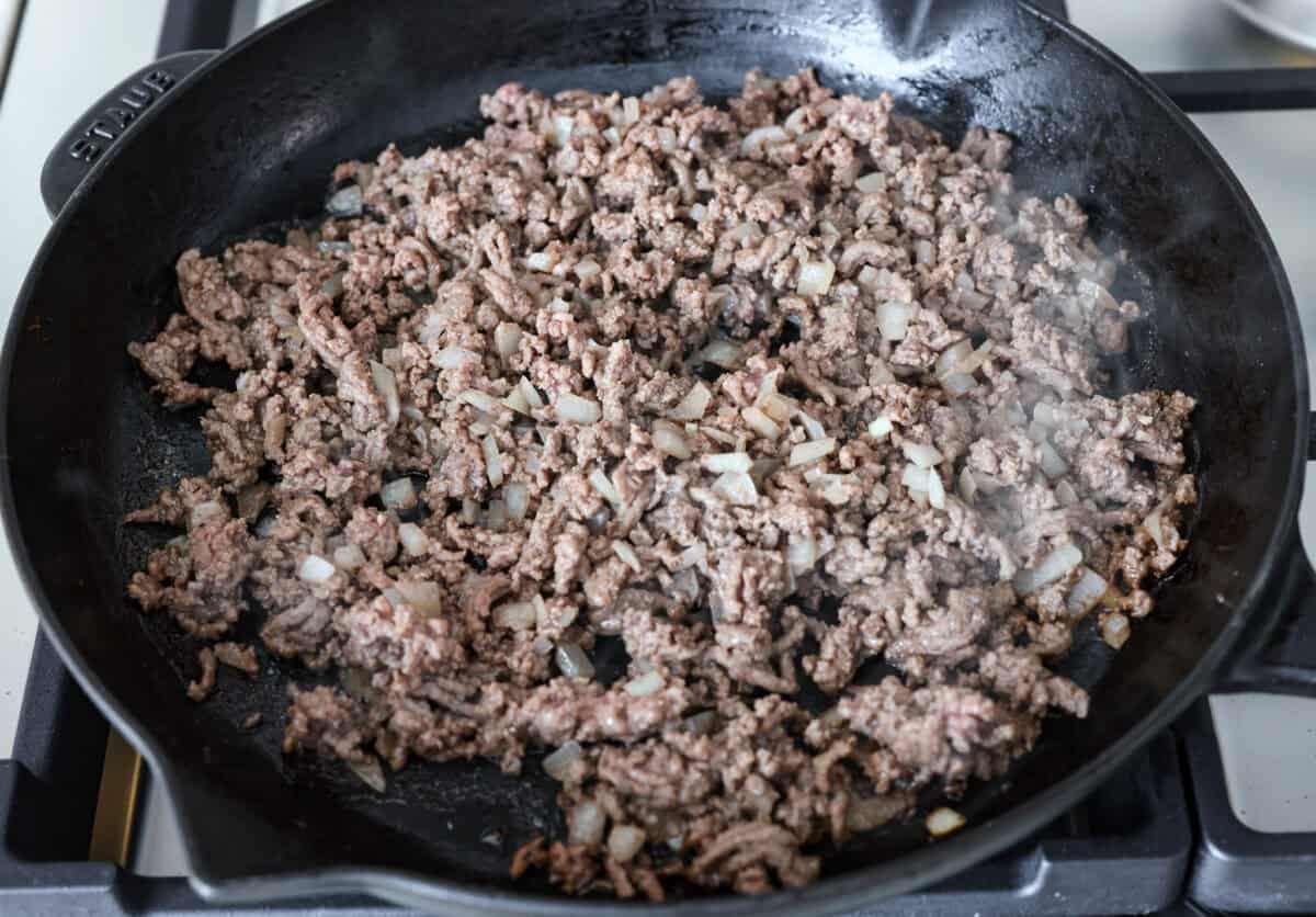This beef enchilada dip packs all of my favorite things into one dish. It starts with red enchilada sauce, savory beef, olives, and sweet corn smothered in melty cheese. Grab your favorite tortilla chips and get dipping. Angle shot of beef and onions sautéing in a skillet.