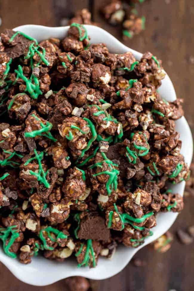 The popcorn is coated in chocolate with Andes Mint pieces and grasshopper mint cookies hidden inside. As soon as I made it I couldn't stop eating it! Overhead photo of Grasshopper Popcorn in a white bowl.
