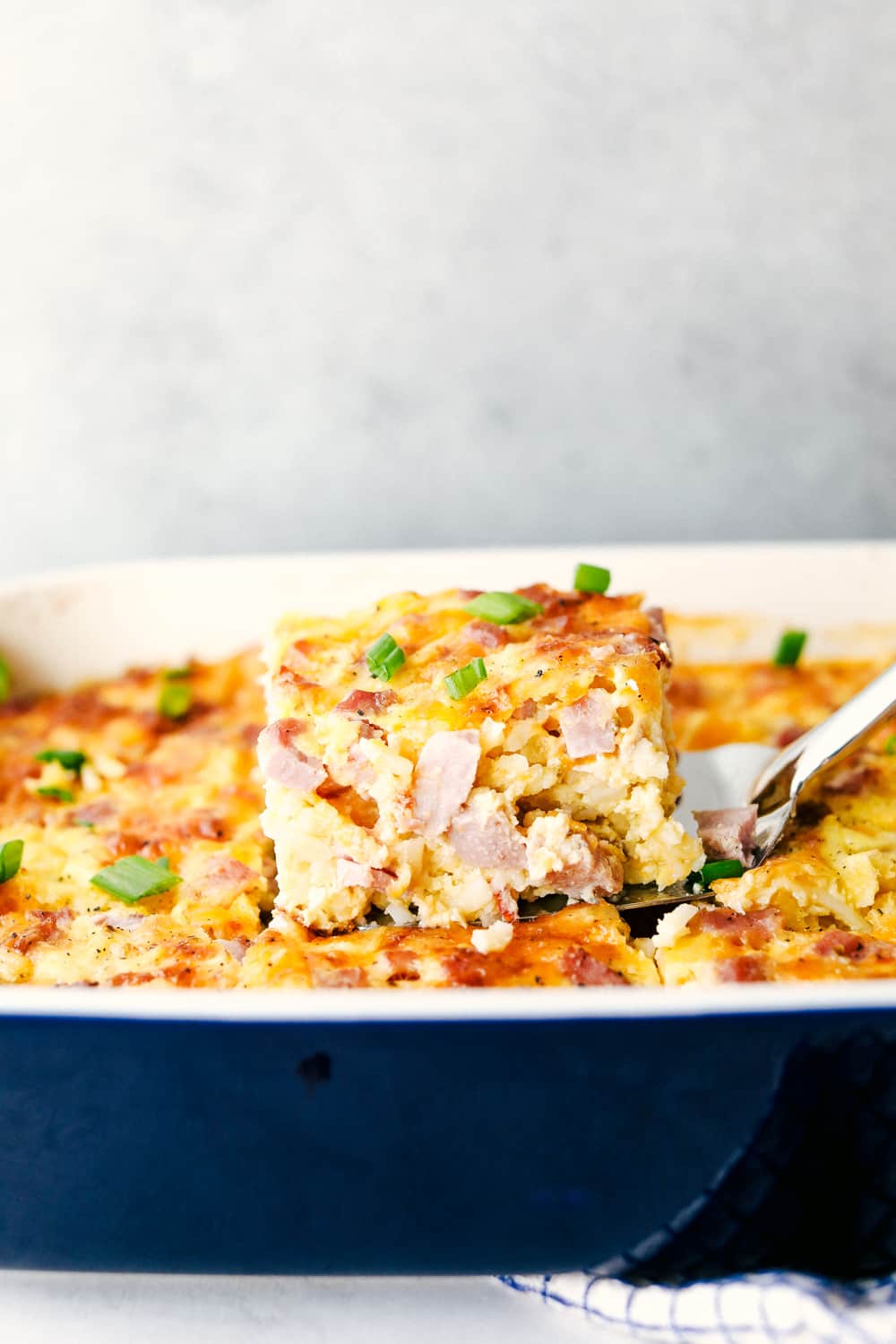 The Best Breakfast Casserole is a thick and creamy egg base with shreddedย potatoes, chunks of ham and flavorful cheese blends that bakes up in no time. Make ahead or enjoy the morning of during the holidays! Side shot of someone lifting out a slice of casserole.