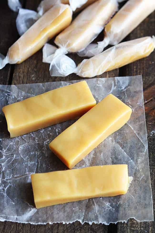 Microwave Caramels are so incredibly easy to make and they turn out perfect! Soft chewy and perfect caramel texture made right in your microwave!ย Unwrapped microwavable Caramels with three of them on a parchment paper with wrapped microwavable Caramels behind them in the photo.