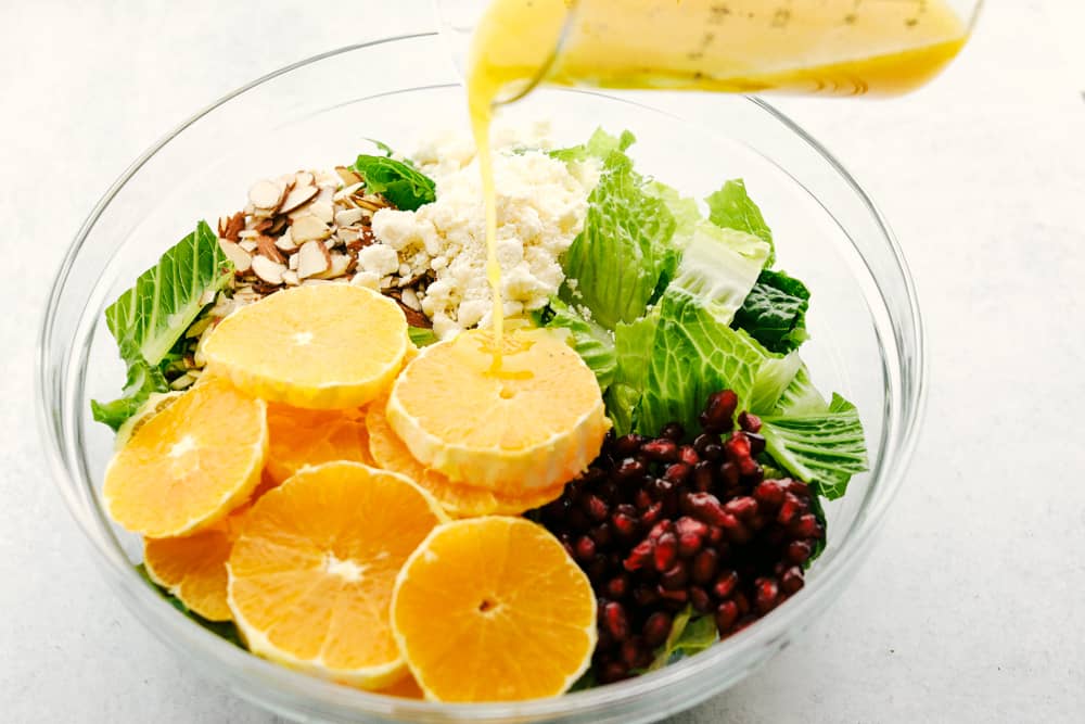 Orange Pomegranate Salad is a delicious mix of greens with fresh oranges, pomegranates and topped with feta cheese and an amazing orange vinaigrette dressing. If you are looking for an awesome salad that is fresh and full of tempting and unique flavors, then this salad is for you! Pouring Orange Vinaigrette dressing over the salad.