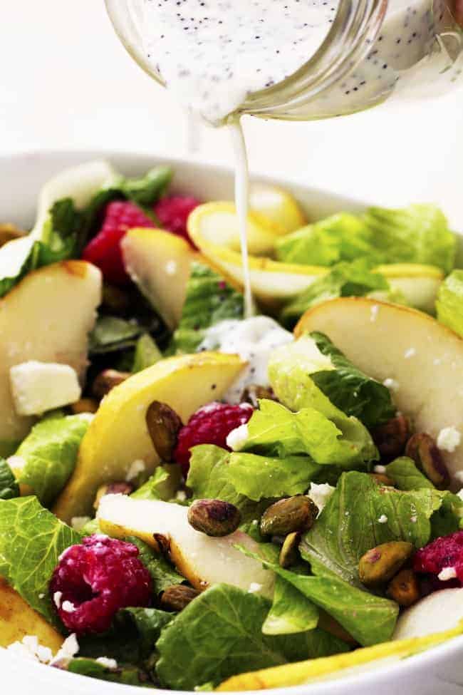 A delicious and healthy salad filled with raspberries, pears, and pistachios add a delicious flavor and crunch! Pouring dressing on pear raspberry pistachio salad.
