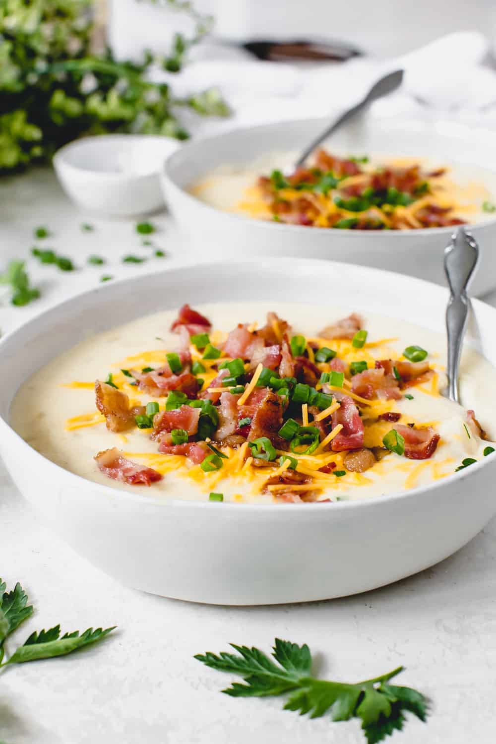 Slow Cooker Loaded Baked Potato Soup is hands down the best loaded baked potato soup that I have ever had. ย Creamy and delicious with all of the goodness of a baked potato and made right in your slow cooker! Two bowls full of loaded baked potato with a spoon garnished with chopped bacon, cheese and green onions.