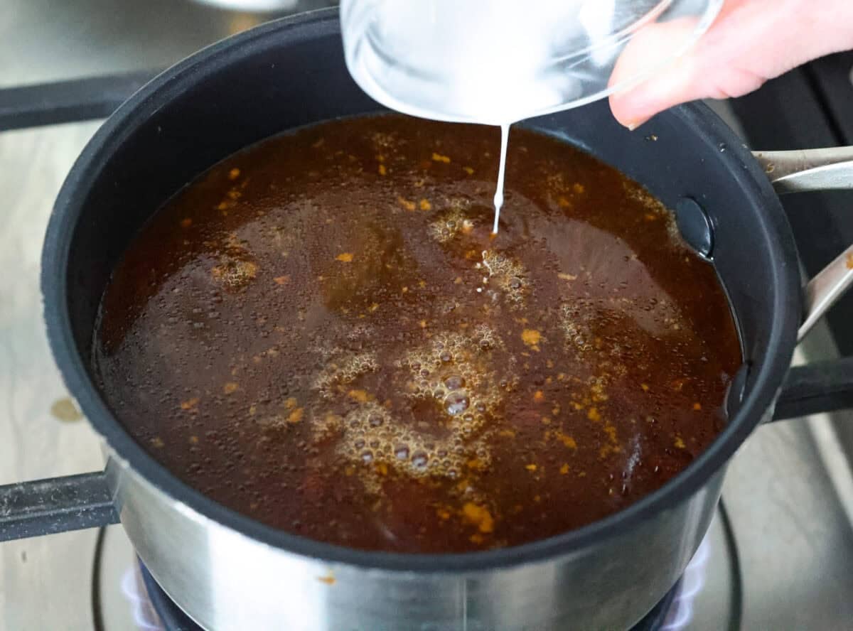 This slow cooker honey teriyaki chicken is so easy to throw together, and the flavor is out of this world! The chicken cooks to perfection and will be one of the best things you'll ever make! Angle shot of someone pouring cornstarch mixture into the sauce in a pot on the stove.