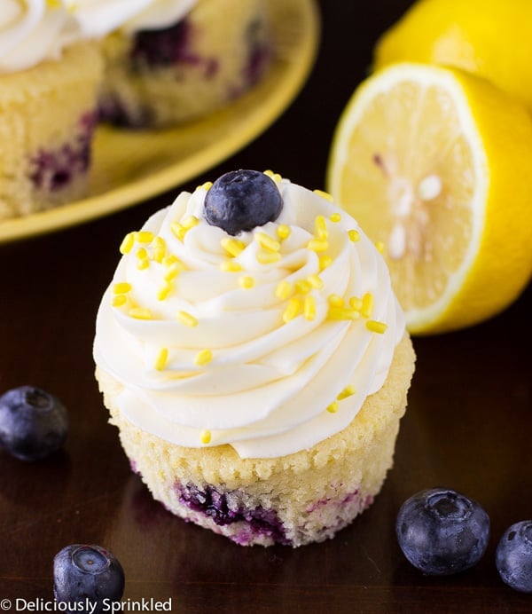 These Lemon Blueberry Cupcakes are bursting with vanilla, fresh blueberries and aย hint of lemon. Piled high with a delicious lemon buttercream frosting and of course, it wouldnโt be a cupcake with out SPRINKLES! Close up on one Lemon Blueberry Cupcake.