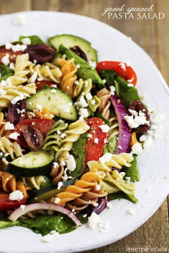 Fresh Spinach combined with olives, red onion, tomatoes, cucumber, and sprinkled with feta cheese. The pasta adds a delicious texture and is tossed with a red wine vinaigrette. Greek spinach pasta salad on a white plate.