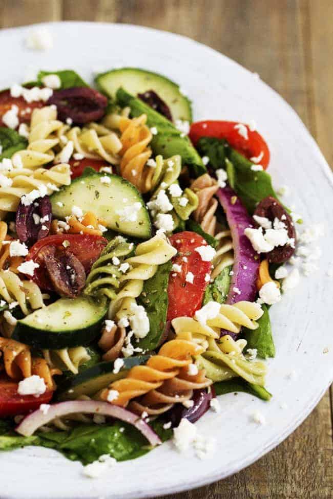 Fresh Spinach combined with olives, red onion, tomatoes, cucumber, and sprinkled with feta cheese. The pasta adds a delicious texture and is tossed with a red wine vinaigrette. greek spinach pasta salad in a white bowl.