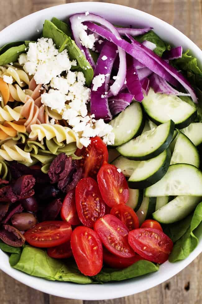 Fresh Spinach combined with olives, red onion, tomatoes, cucumber, and sprinkled with feta cheese. The pasta adds a delicious texture and is tossed with a red wine vinaigrette. greek spinach pasta salad ingredients in a white bowl.