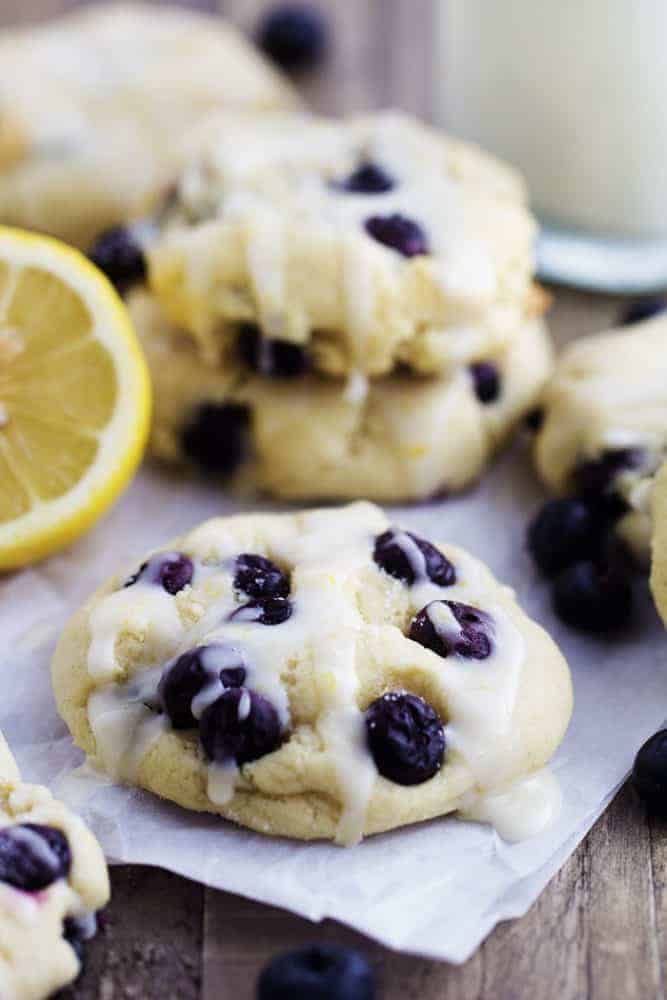 Perfect moist and puffy cookies with fresh blueberries bursting inside. These cookies are a mix between a blueberry muffin and a soft and chewy cookie. Drizzled in a lemon glaze, these will become a new favorite! Blueberry cream cheese cookies with a glaze resting on a white paper.