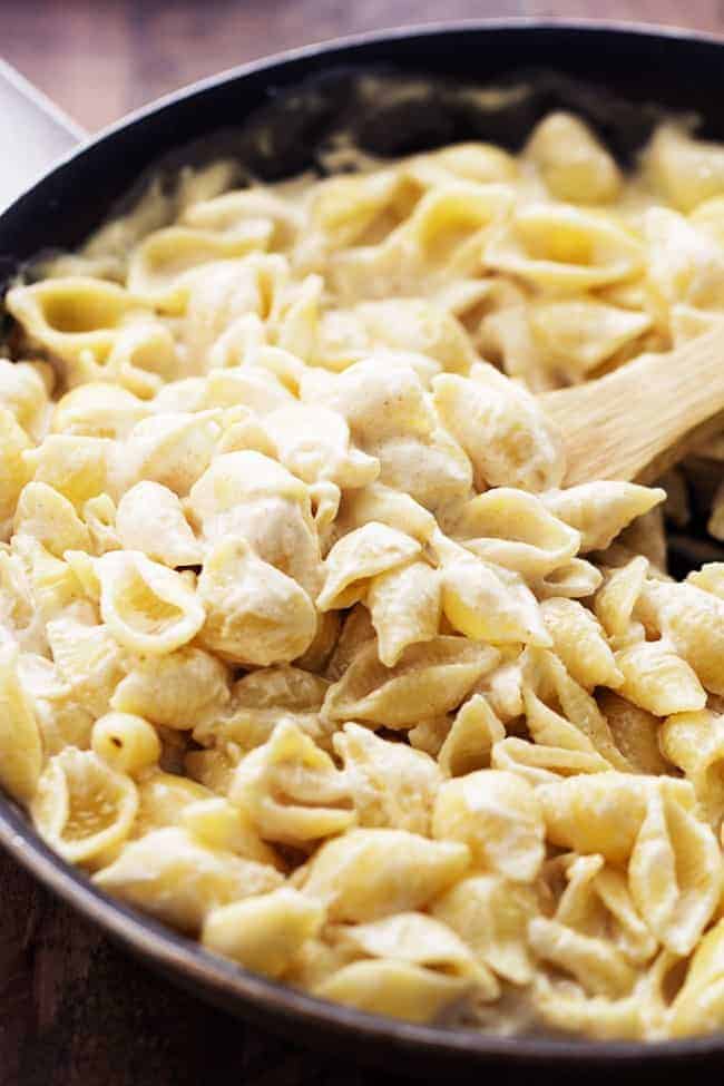 The most creamy and delicious homemade garlic alfredo shells topped with crispy parmesan garlic chicken. This is an absolutely fantastic meal that will instantly become a family favorite! Wooden spoon stirring creamy shells in a pan.