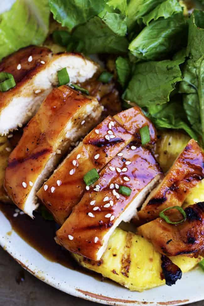 The most delicious sweet pineapple teriyaki glaze coats this chicken perfectly. It grills to juicy and tender perfection on the BBQ and will be one of the best things you grill this summer! Grilled pineapple teriyaki chicken with salad and pineapple on a white plate.