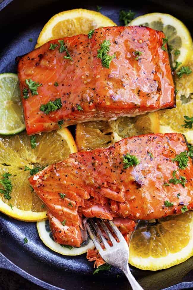 A five starย salmon recipe that gets infused with a lemon, lime and orange marinade. It grills to tender and flaky perfection and has the best sweet and tangy flavor! grilled citrus salmon on lemon slices in a skillet being cut into by a fork.