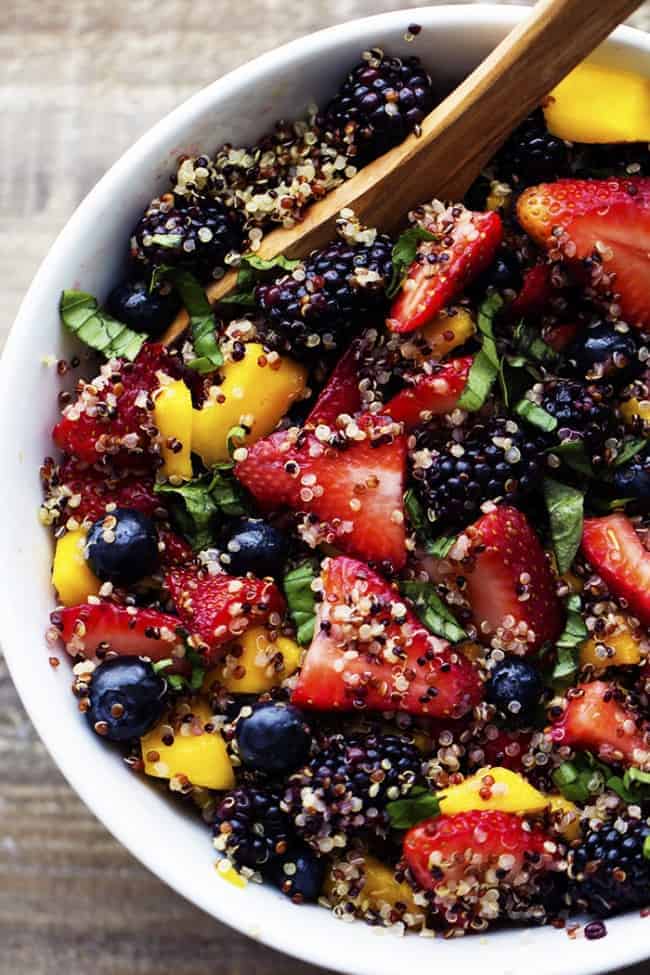 Quinoa, strawberries, blueberries, blackberries, and mango combine in this incredible salad. The fresh fruit and quinoa are glazed with a honey lime dressing and it will be a hit wherever it goes! honey lime quinoa fruit salad in a white bowl with a wooden spoon.
