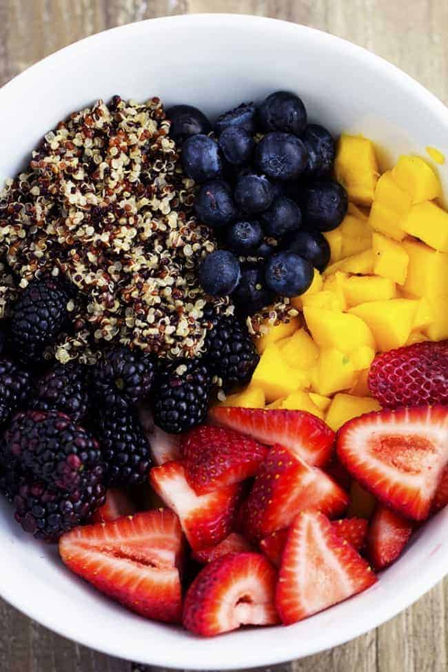 Quinoa, strawberries, blueberries, blackberries, and mango combine in this incredible salad. The fresh fruit and quinoa are glazed with a honey lime dressing and it will be a hit wherever it goes! honey lime quinoa salad ingredients in a white bowl.