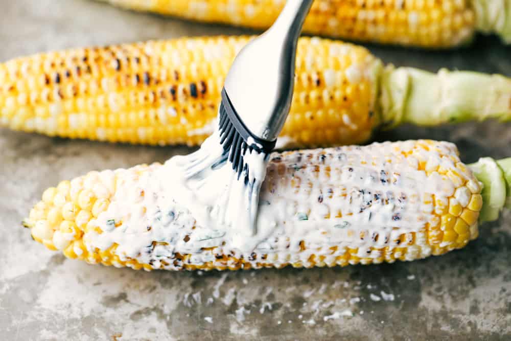 This corn gets grilled and slathered with an amazing mayonnaise blend and topped with parmesan cheese and chili powder. This is one of the best corn recipes you will eat! Grilled corn being brushed with mayonnaise.