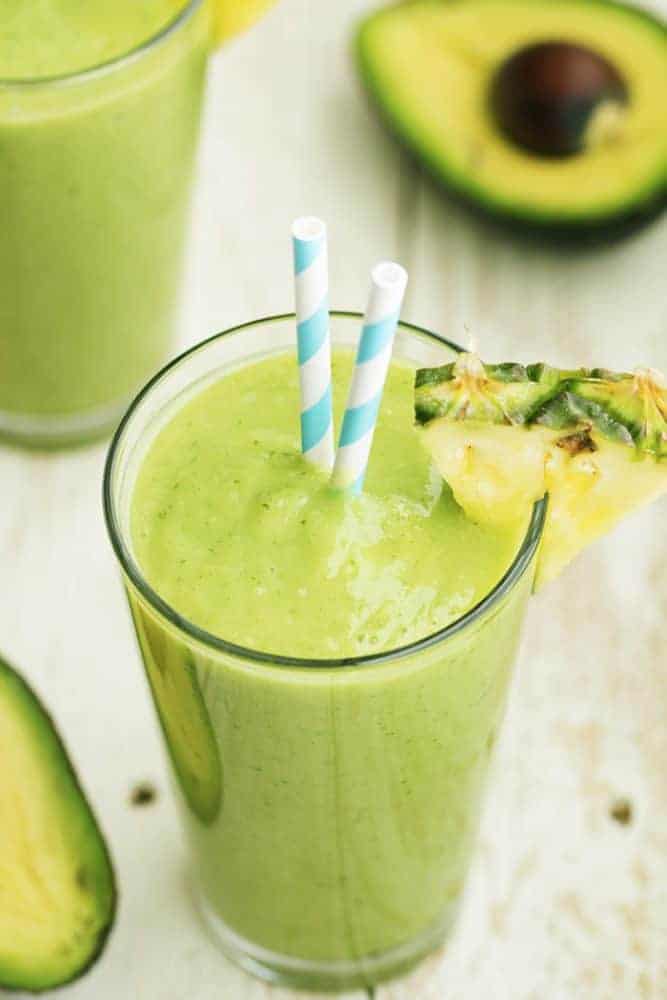 This delicious and perfectly smooth avocolada smoothie combines avocado, pineapple, spinach, kale, coconut, and lime. It's SOO GOOD that you will want one every day! Avocolada smoothie in a glass with pineapple garnish and blue and white striped straws.