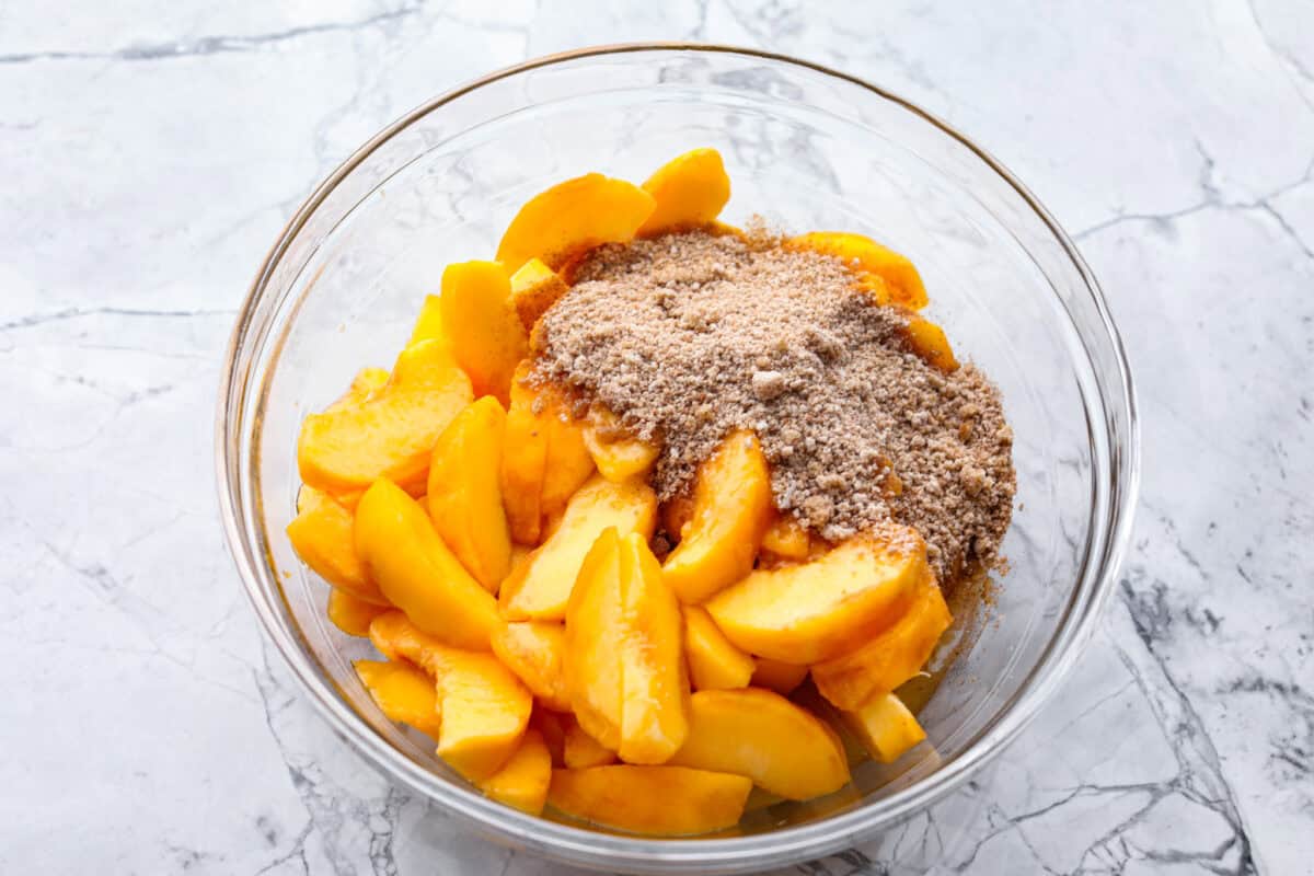 Old-fashioned peach cobbler is a timeless summer dessert! Its warm, cakey layers baked with freshly sliced peaches are a match made in heaven. Top the cobbler with vanilla ice cream, and every bite will be a delight! Overhead shot of fresh sliced peaches with cinnamon sugar mixture in glass bowl.