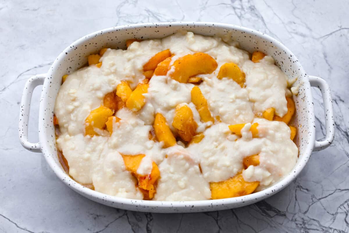 Old-fashioned peach cobbler is a timeless summer dessert! Its warm, cakey layers baked with freshly sliced peaches are a match made in heaven. Top the cobbler with vanilla ice cream, and every bite will be a delight! Overhead shot of old fashioned peach cobbler mixture topped with biscuit topping in baking dish, ready to be baked.