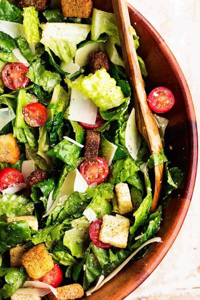 Fresh romaine and cherry tomatoes get tossed with a delicious asiago caesar dressing. Topped with shaved parmesan cheese and croutons, this is a delicious and classic salad that makes the perfect side! classic caesar salad areal view in a red bowl with wooden spoon.