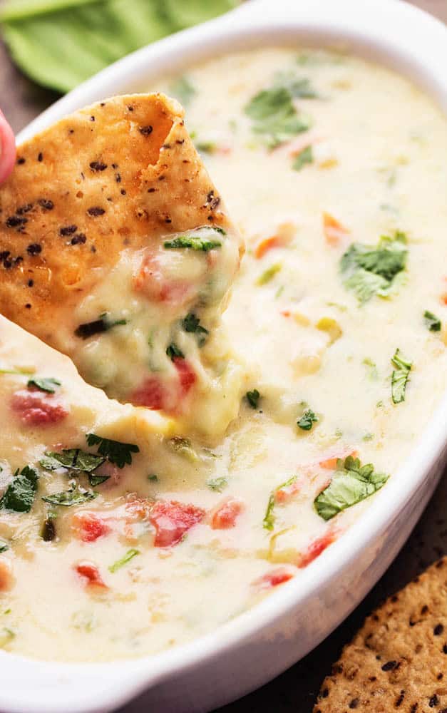 Melty white cheese, spinach, jalapeรฑos, and diced tomatoes come together in this incredible dip! This is a must make dip that you won't be able to get enough of! White Spinach Queso in a white dish with a tortilla chip dipping into it.