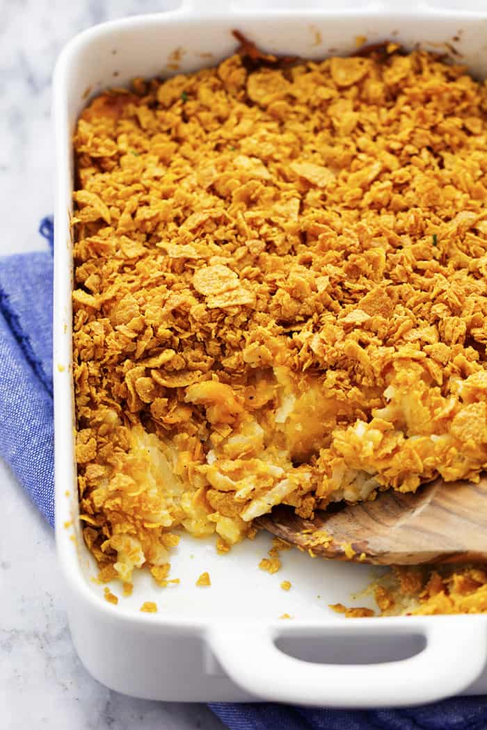 A cheesy potato casserole full of melted cheese, sour cream, onions, and garlic with a crunchy top. This is full of amazing flavor and will be the star of the dinner table! funeral potatoes in a white baking dish with wooden spoon scooping out potatoes.