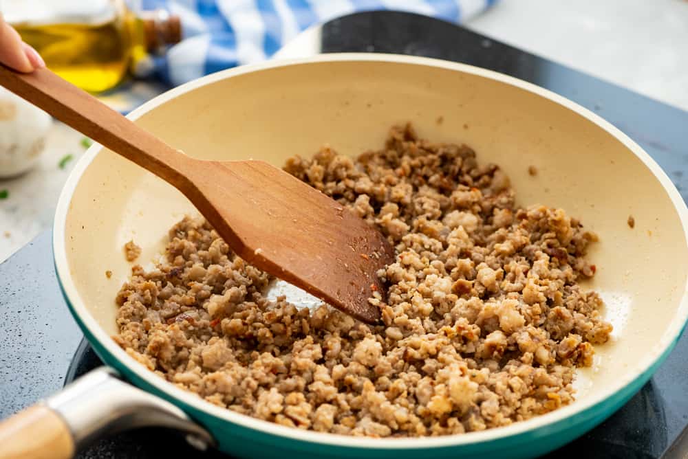 Sausage Stuffed Mushrooms are stuffed mushroom caps filled with a creamy and delicious mixture of onions, garlic, chopped mushroom stems, and sausage! These are a must-make for your next party! Sausage being cooked in a blue and white skillet.