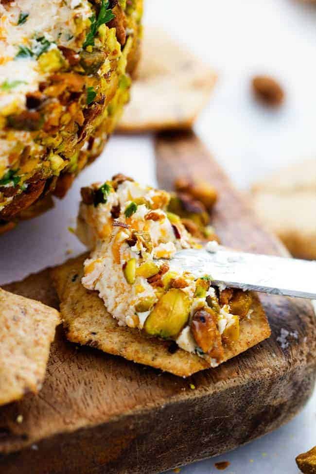 An amazing cheeseball packed with all of my favorite things! ย Cheddar, pistachios, and bacon combine to make one delicious cheeseball that is perfect for your next party!ย Cheddar Bacon Pistachio Cheeseball with being spread on cracker