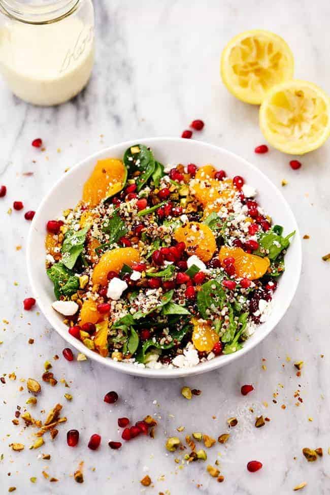 A mouthwatering winter salad with quinoa, spinach, mandarine oranges, and crunchy pistachios. Topped with a creamy lemon greek yogurt dressing and feta cheese this makes one unforgettable salad! WINTER POMEGRANATE ORANGE QUINOA SALAD WITH CREAMY LEMON GREEK YOGURT DRESSING IN A WHITE BOWL WITH TWO FRESHLY SQUEEZED LEMONS ON THE SIDE.