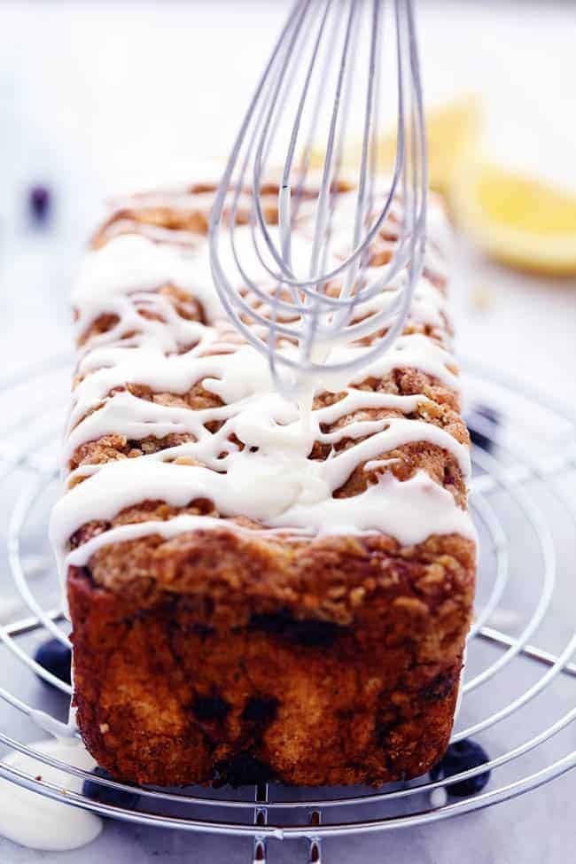 An amazingly moist muffin bread filled with blueberry pie filling. Topped with a crumb topping and drizzled in a lemon glaze this bread is incredible!ย Blueberry pie bread on a cooling rack with lemon glaze being spread by a whisk.