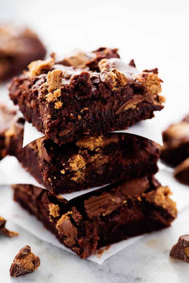 Ultra rich brownies with a creamy hot fudge cheesecake centerย and topped with peanut butter cups. So many delicious things in one amazing brownie that your family will go crazy for! Hot Fudge Peanut Butter Cup Cheesecake Brownies stacked on top of one another with parchment paper in between them.