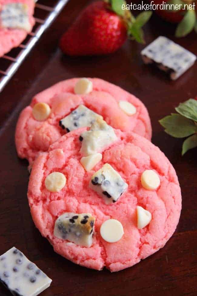 Easy and delicious cookies that are made with a cake mix and pudding mix with delicious cookies and cream pieces hidden inside! They are a fun pink color with a crisp edge and chewy center! Strawberry Cookies 'n Cream Cookies on table with fresh strawberries in background.
