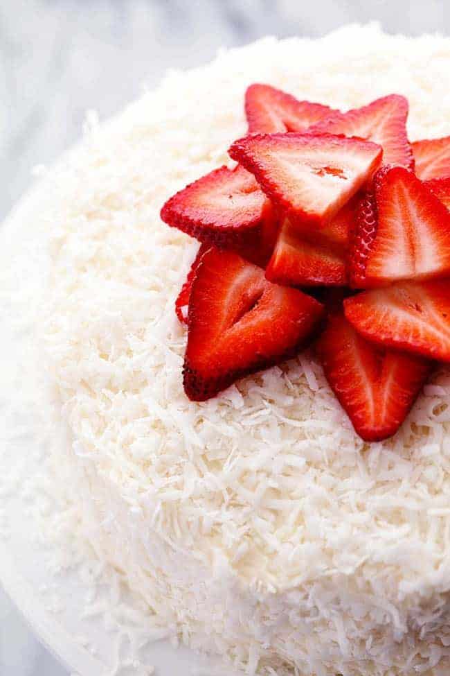 A delicious and moist strawberry coconut cream cake with an amazing coconut cream cheese frosting! Strawberry Coconut Cream Cake with Coconut Cream Cheese Frosting with fresh cut strawberries on a white cake stand.