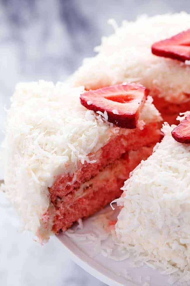 A delicious and moist strawberry coconut cream cake with an amazing coconut cream cheese frosting! Strawberry Coconut Cream Cake with Coconut Cream Cheese Frosting on a white cake stand with a slice being removed.