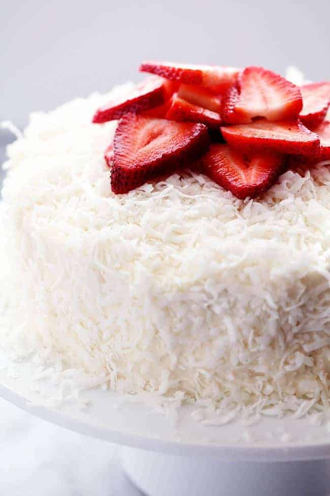 A delicious and moist strawberry coconut cream cake with an amazing coconut cream cheese frosting! Strawberry Coconut Cream Cake with Coconut Cream Cheese Frosting and fresh cut strawberries on top displayed on a white cake stand.
