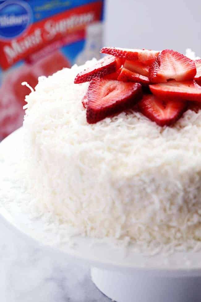 A delicious and moist strawberry coconut cream cake with an amazing coconut cream cheese frosting! Strawberry Coconut Cream Cake with Coconut Cream Cheese Frosting with fresh cut strawberries on a white cake stand.