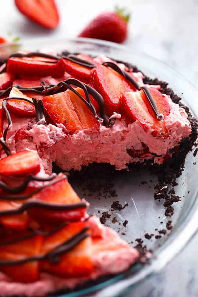 An amazing no bake pie with a chocolate crust filled with a creamy strawberry filling. It gets topped with fresh strawberries and drizzled in chocolate and will be a huge hit wherever it goes!ย No Bake Chocolate Strawberries and Cream Pie with a slice removed in a clear pie pan.