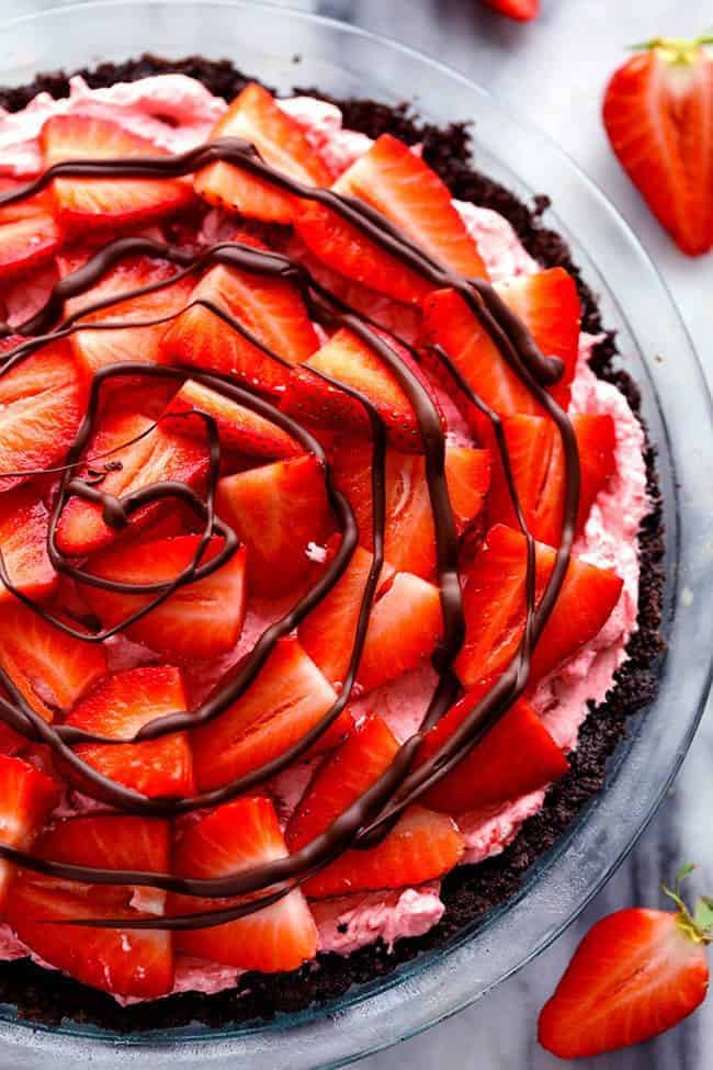 An amazing no bake pie with a chocolate crust filled with a creamy strawberry filling. It gets topped with fresh strawberries and drizzled in chocolate and will be a huge hit wherever it goes!ย Overhead photo of No Bake Chocolate Strawberries and Cream Pie in a clear pie dish.