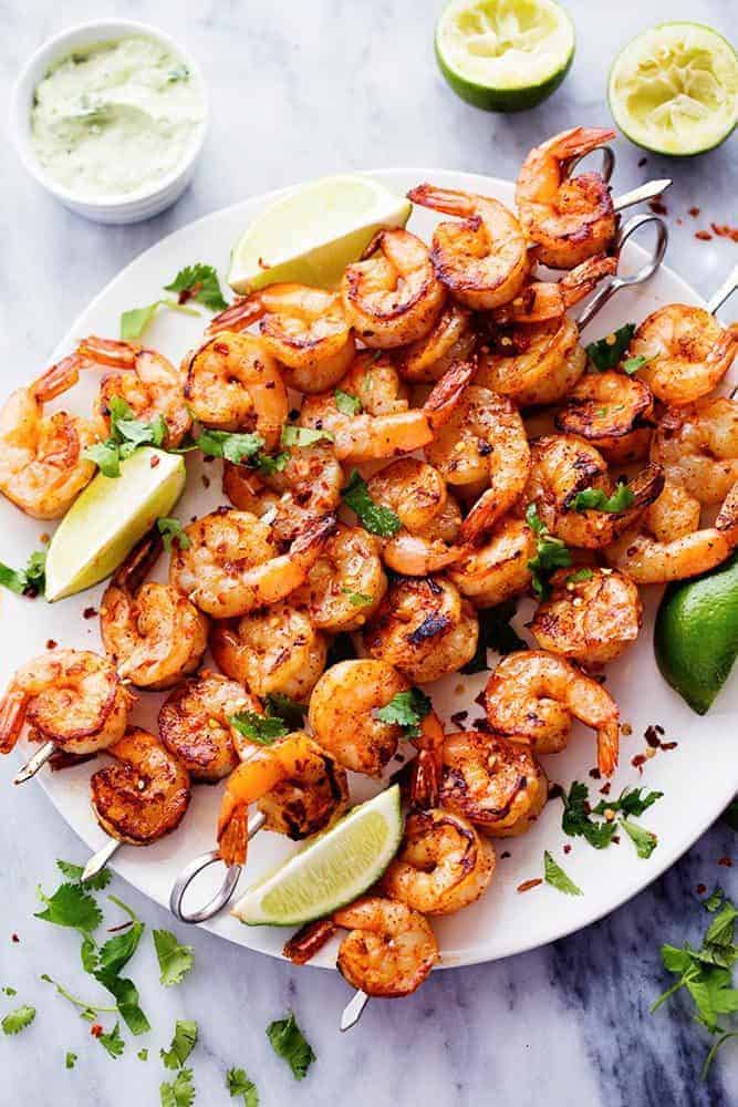 Grilled Spicy Lime Shrimp with Creamy Avocado Cilantro Sauce has a simple but full of flavor and spice marinade. The creamy avocado cilantro sauce is the perfect cool and creamy dipping sauce. Grilled Spicy Lime Shrimp with Creamy Avocado Cilantro Sauce on skewers on a white plate with fresh limes on the side.