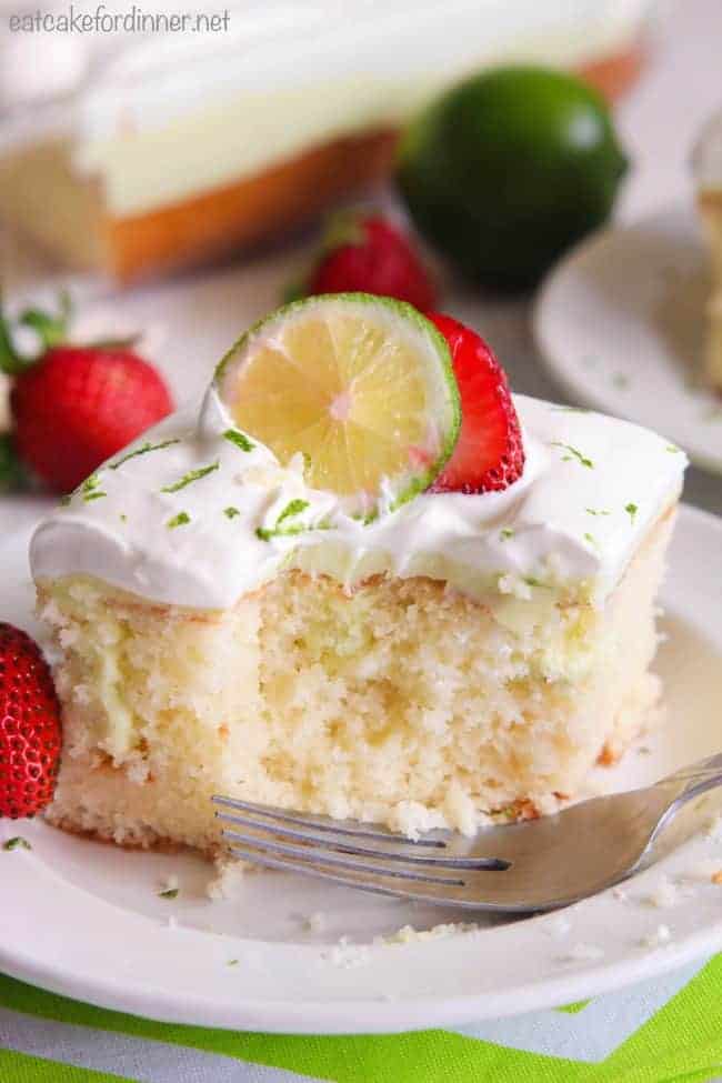 Key Lime Poke Cake is a delicious white cake that is seeping with a sweet and tart mixture that soaks into the holes. Topped with fresh whipping cream and strawberries this makes a delicious dessert! Key Lime Poke Cake on a white plate with fresh strawberries and lime on top.