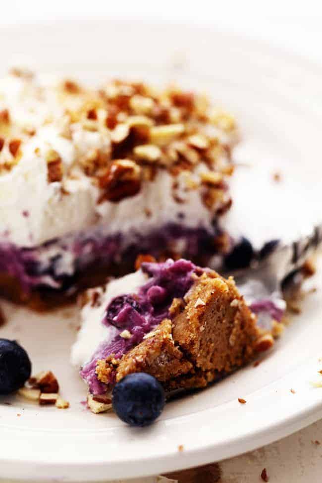 No Bake Creamy Blueberry Pecan Squares are a cool and creamy layered summer dessert. You will love the creamy blueberry center on top of a pecan graham crust and topped with fresh whipping cream and chopped pecans! No Bake Creamy Blueberry Pecan Square on a white plate.