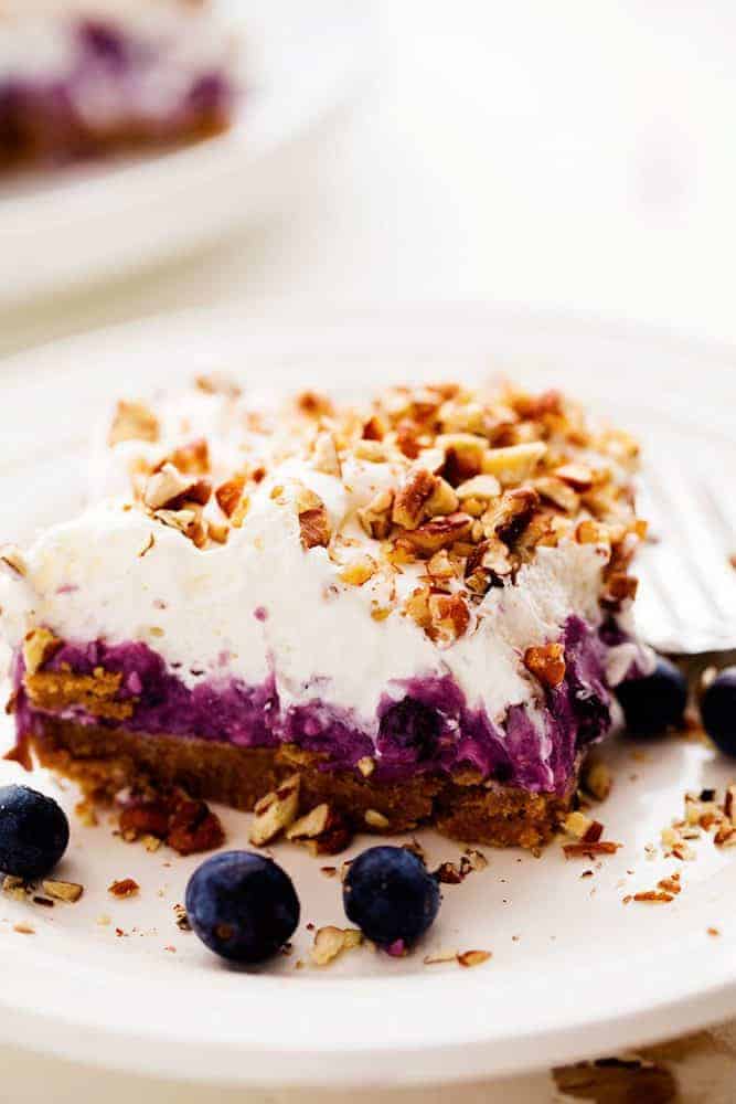 No Bake Creamy Blueberry Pecan Squares are a cool and creamy layered summer dessert. You will love the creamy blueberry center on top of a pecan graham crust and topped with fresh whipping cream and chopped pecans! No Bake Creamy Blueberry Pecan Square on a white plate with a metal fork and fresh blueberries on the side.
