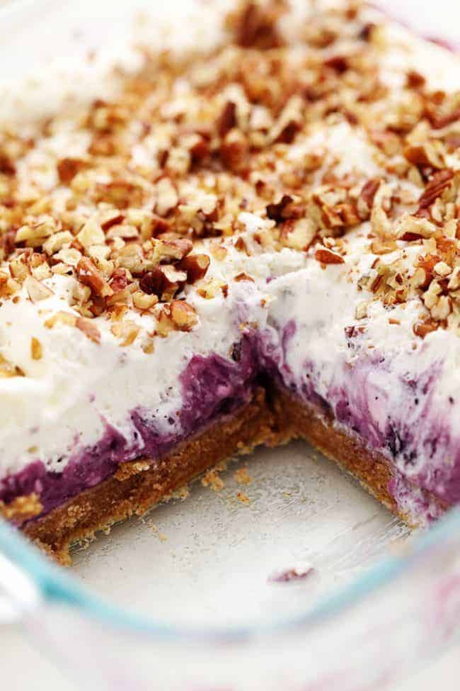 No Bake Creamy Blueberry Pecan Squares are a cool and creamy layered summer dessert. You will love the creamy blueberry center on top of a pecan graham crust and topped with fresh whipping cream and chopped pecans! No Bake Creamy Blueberry Pecan Squares in a clear pan with one square removed.