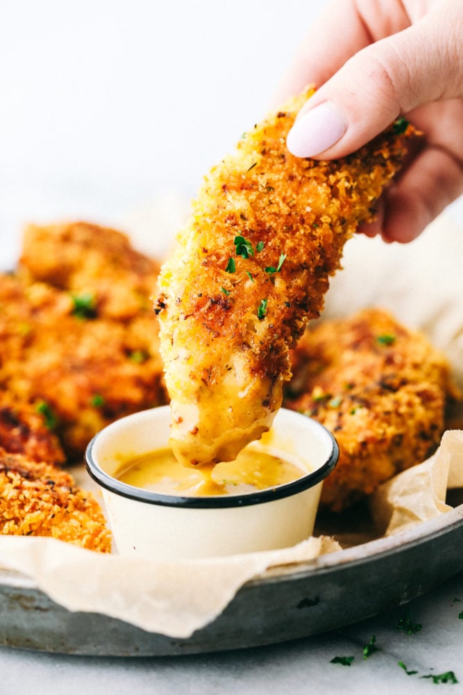 Delicious chicken tenders breaded in a garlic parmesan panko mixture and baked to crispy perfection! This is a meal the entire family will love! Chicken tender being dipped in a honey mustard sauce.