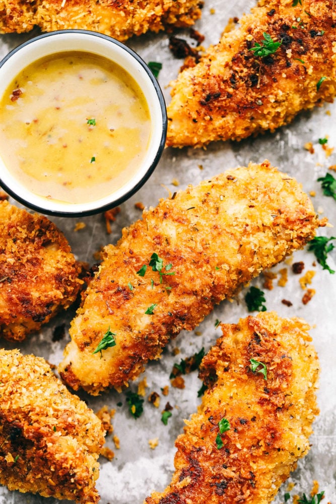 Delicious chicken tenders breaded in a garlic parmesan panko mixture and baked to crispy perfection! This is a meal the entire family will love! Chicken tenders on a baking sheet with honey mustard sauce.