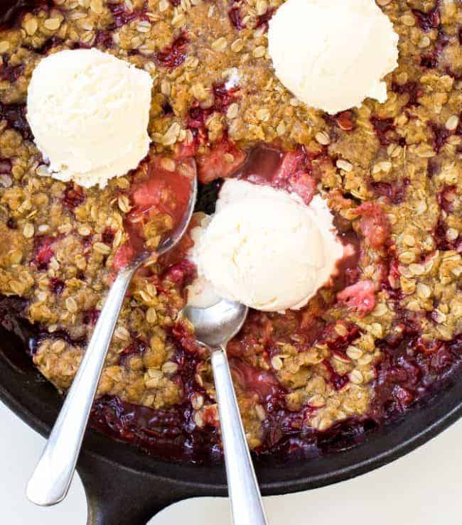 Super Easy Strawberry Crisp. The perfect dessert for summer! Strawberry Crisp in a large black skillet with three scoops of ice cream and two metal spoons.