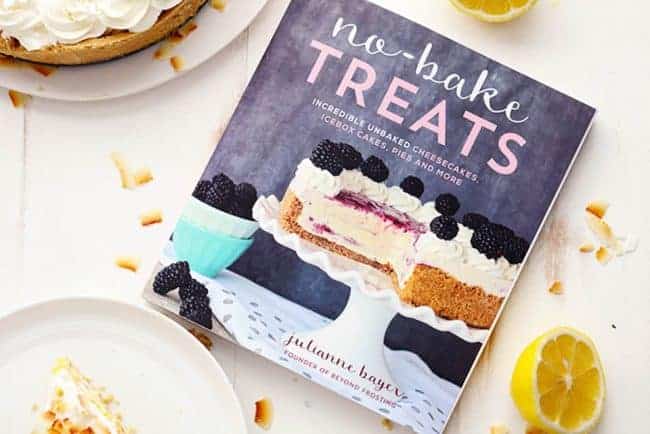 No Bake Lemon Macaroon Cheesecake is a coconut filled cheesecake on top of a golden oreo crust. Topped with lemon curd andย fresh whipping cream this easy to make dessert will be a huge hit! Cookbook named "No-Bake Treats" with a couple of white plates next to it.