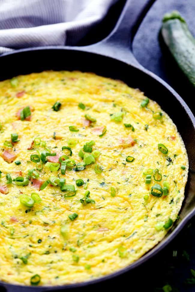 Ham and Cheddar Zucchini Frittata is filled with two cups of zucchini, ham, and cheddar cheese. It has such amazing flavor and will be devoured in no time at all! Ham and Cheddar Zucchini Frittata in a large pan with a Zucchini on the side.
