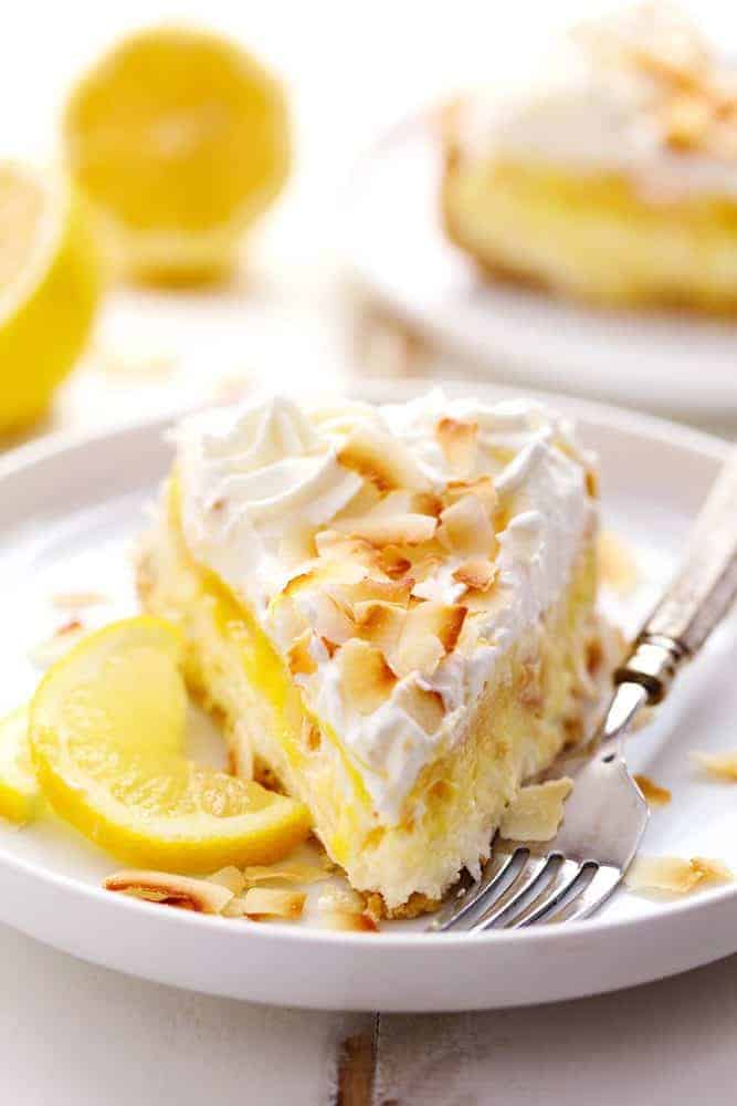 No Bake Lemon Macaroon Cheesecake is a coconut filled cheesecake on top of a golden oreo crust. Topped with lemon curd andย fresh whipping cream this easy to make dessert will be a huge hit! A slice of No Bake Lemon Macaroon Cheesecake on a white plate with a metal fork. There is fresh sliced lemon in the background.