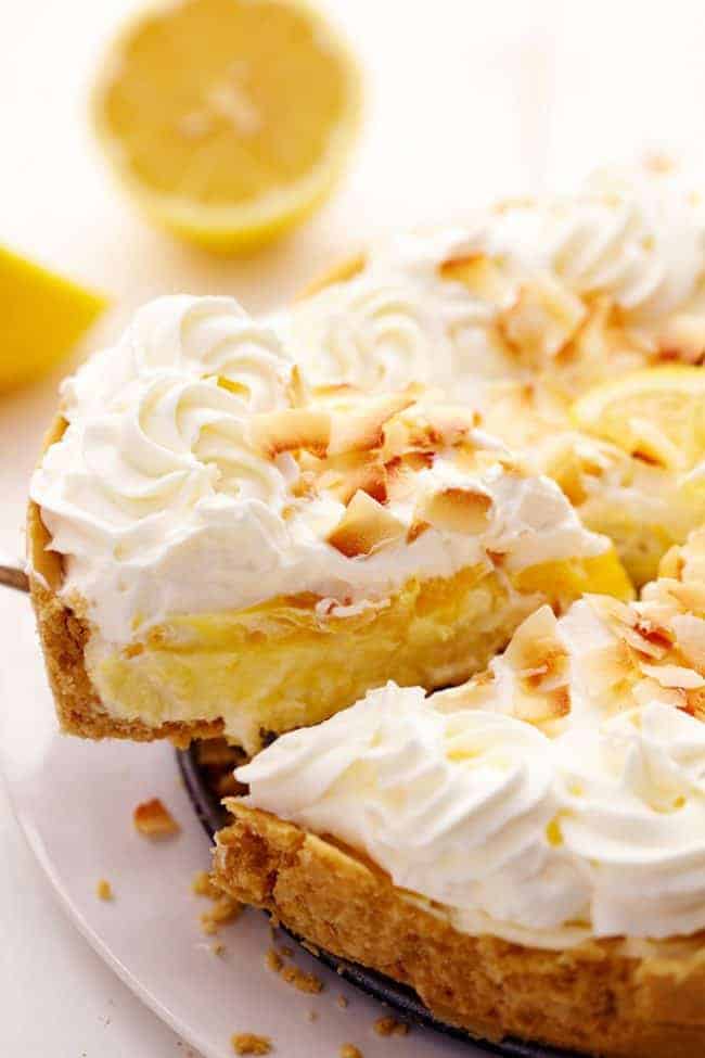 No Bake Lemon Macaroon Cheesecake is a coconut filled cheesecake on top of a golden oreo crust. Topped with lemon curd andย fresh whipping cream this easy to make dessert will be a huge hit! No Bake Lemon Macaroon Cheesecake with a slice being removed and lemons in the background.