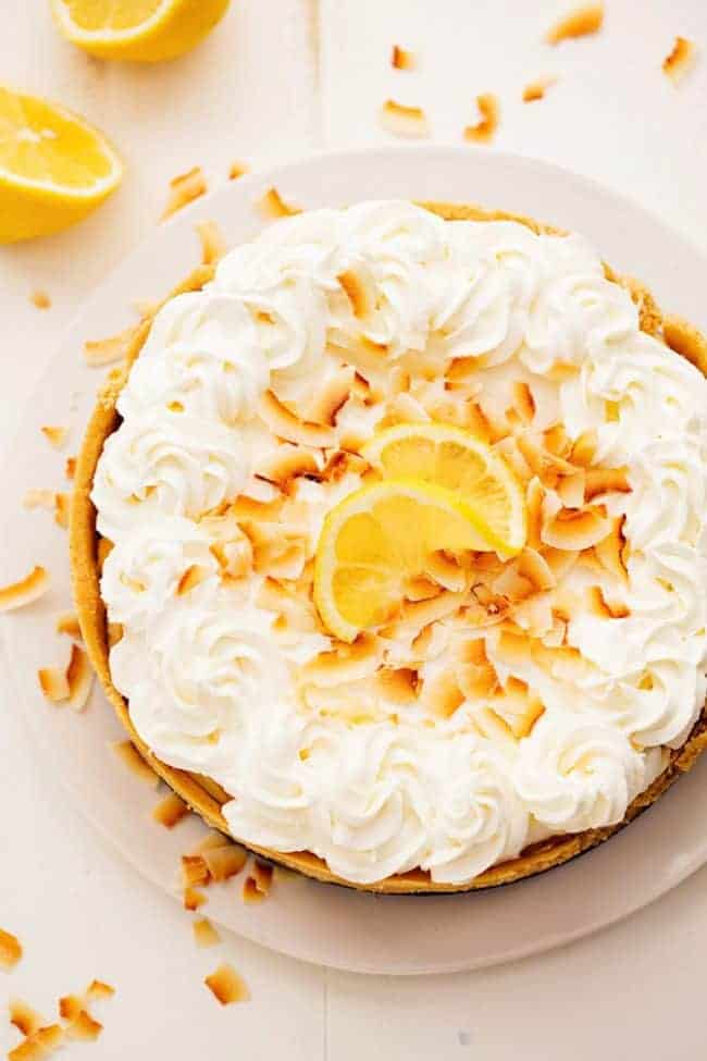 No Bake Lemon Macaroon Cheesecake is a coconut filled cheesecake on top of a golden oreo crust. Topped with lemon curd andย fresh whipping cream this easy to make dessert will be a huge hit! No Bake Lemon Macaroon Cheesecake on a white plate with fresh lemons on the side.